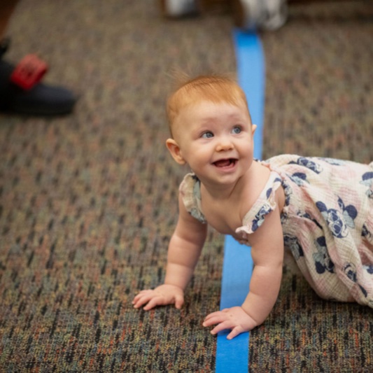 A smiling baby crawling along a taped racetrack line on the library carpet