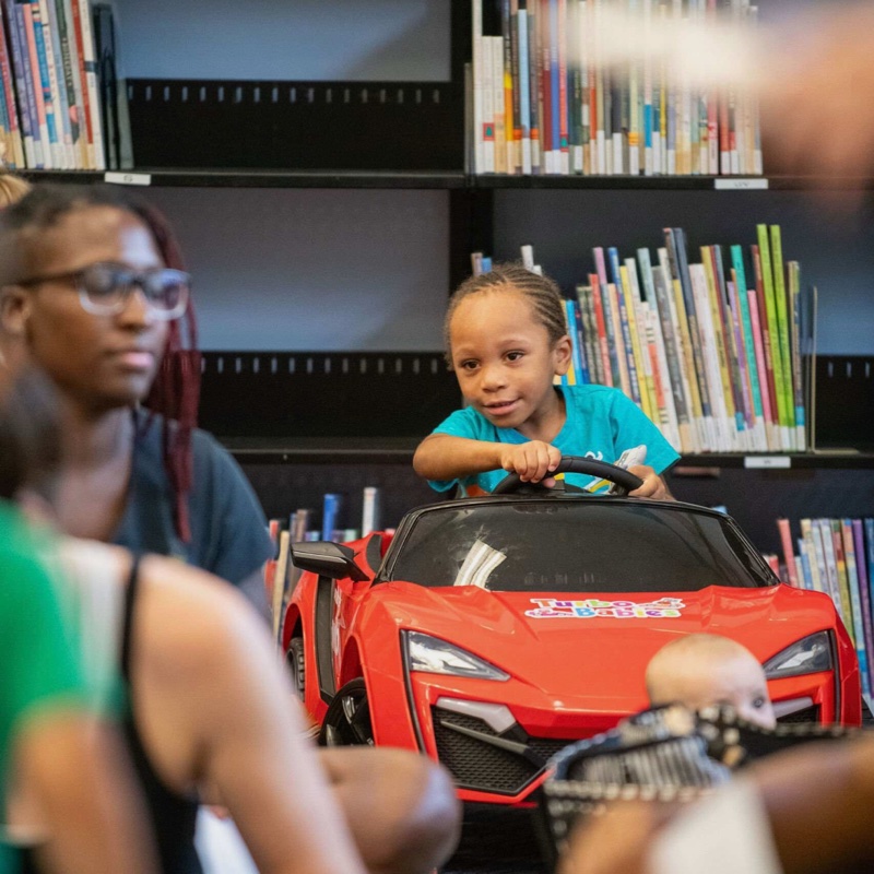 A young child steering a red Turbo Babies ride-on car at the library